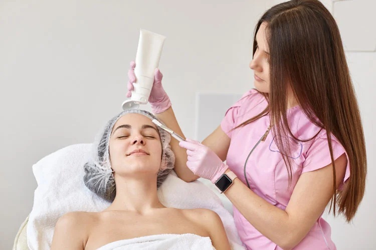 Médecin cosmétologue effectuant un massage liftant du visage d'une jeune femme brune, cliente allongée sur le divan, les yeux fermés, portant une serviette blanche et une casquette médicale.